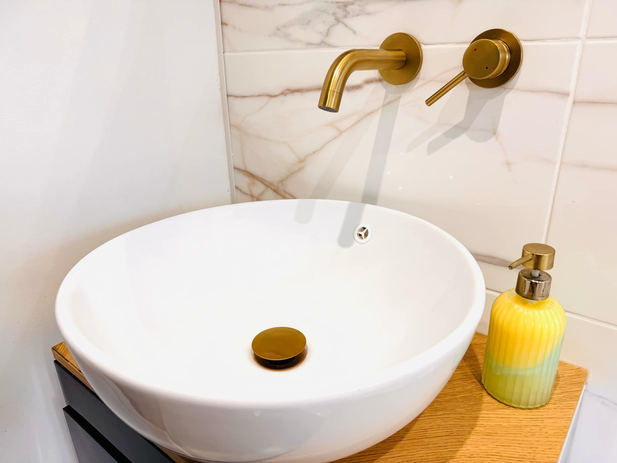 Un lavandino in ceramica bianca con un rubinetto dorato e un dispenser di sapone giallo su un piano di lavoro in legno.