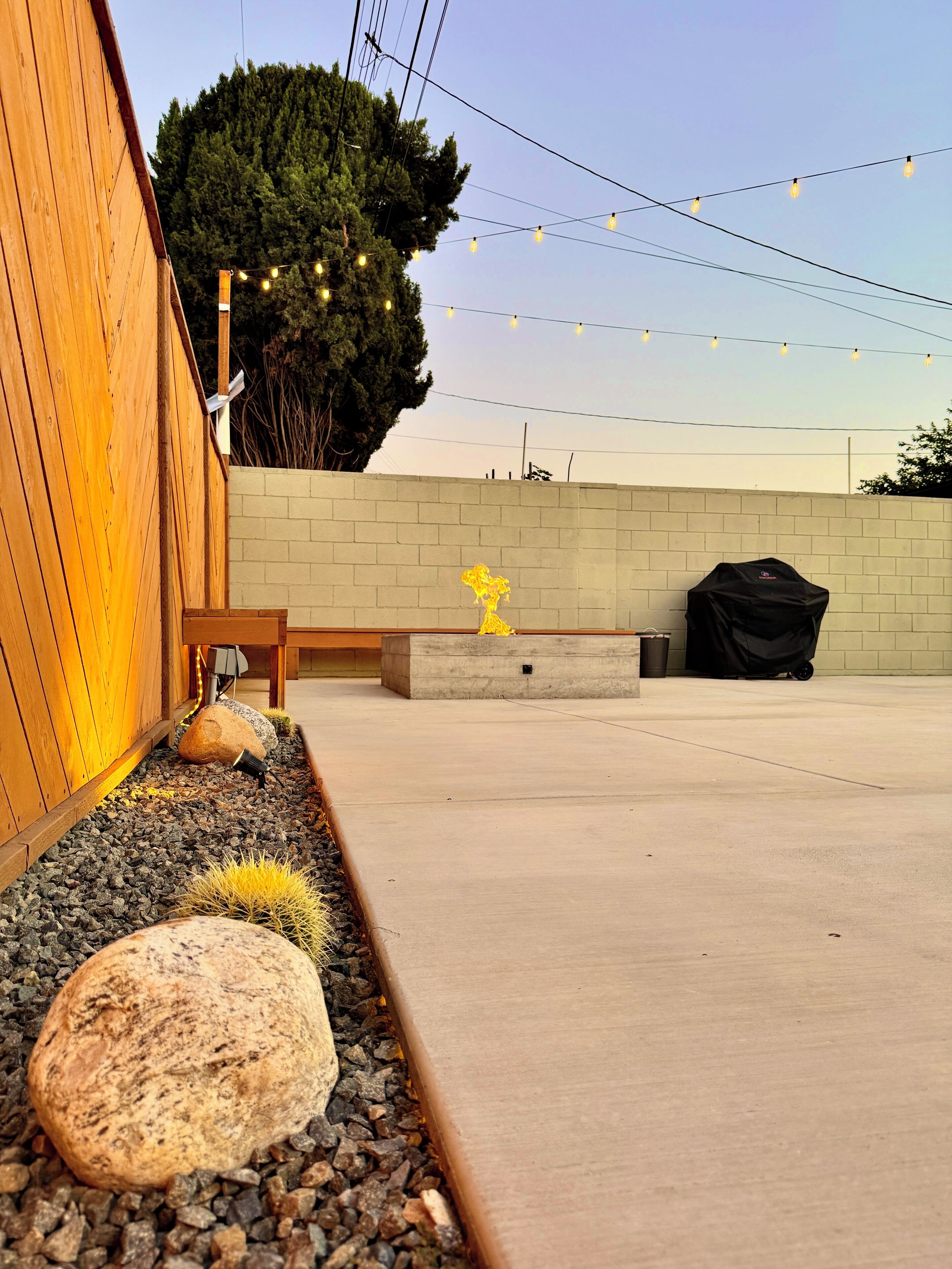 An outdoor patio area with a concrete floor, a wooden fence, a stone planter with cacti, and a barbecue grill.