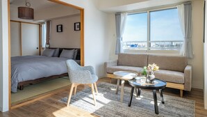 A modern hotel room with a bed, a sofa, a small table with a vase of flowers, and a view of the city through the window. - E-US NAEBO601 (Sapporo)