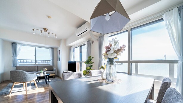 A modern living room with a dining table, sofa, and chairs. There is a large window with curtains, a vase with flowers, and an air conditioning unit on the wall. - E-US NAEBO601 (Sapporo)