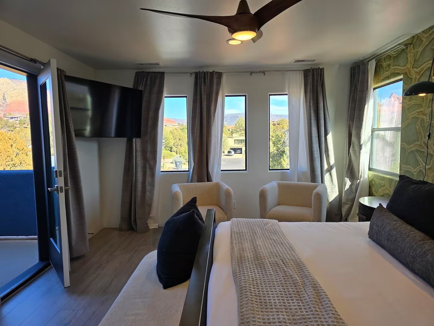 Un dormitorio moderno con cama, dos sillones, televisor y ventanas amplias con vistas al exterior.