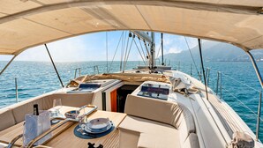 Cubierta de barco con zona de estar cubierta, mesa de comedor y vista al mar.