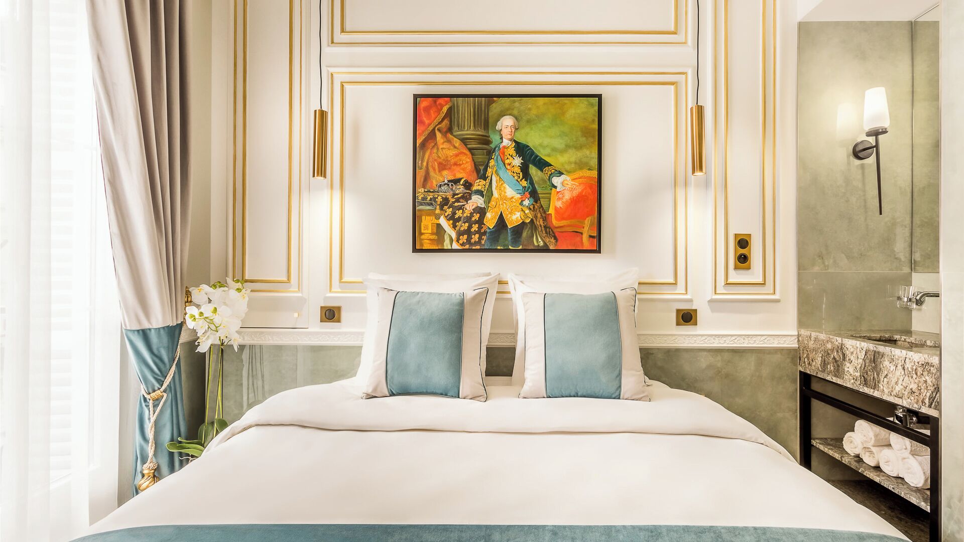 Une chambre d’hôtel avec un grand lit, un tableau au-dessus du lit, une salle de bain avec un miroir et un porte-serviettes, et un vase avec des fleurs sur le côté gauche.
