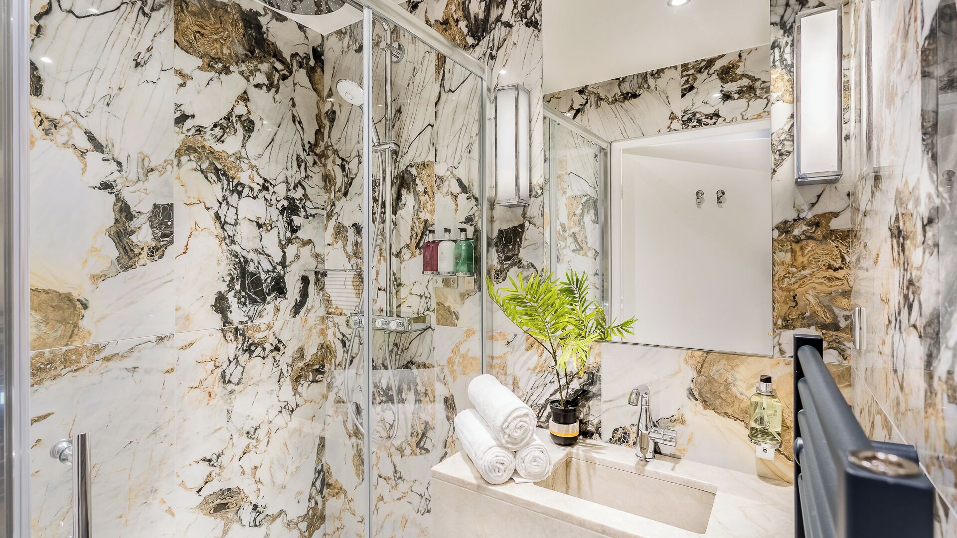 A bathroom with marble walls, a glass-enclosed shower, a sink with a mirror, and a potted plant.
