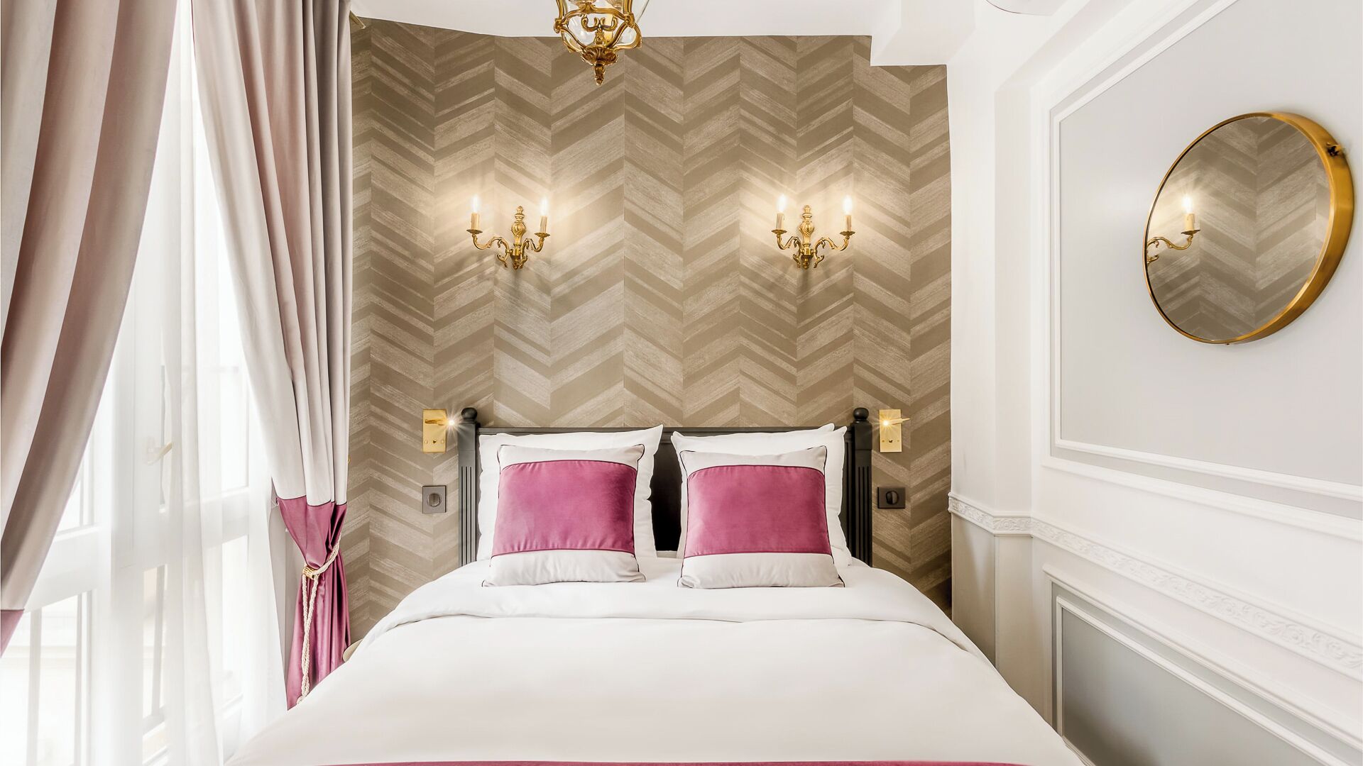 Habitación de hotel con cama, dos apliques, un espejo redondo y un papel tapiz con estampado en zigzag.