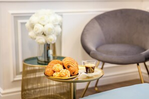 Egy asztal, rajta croissant-okkal és egy csésze kávéval, egy fehér virágokkal teli váza, és egy szürke színű karosszék.