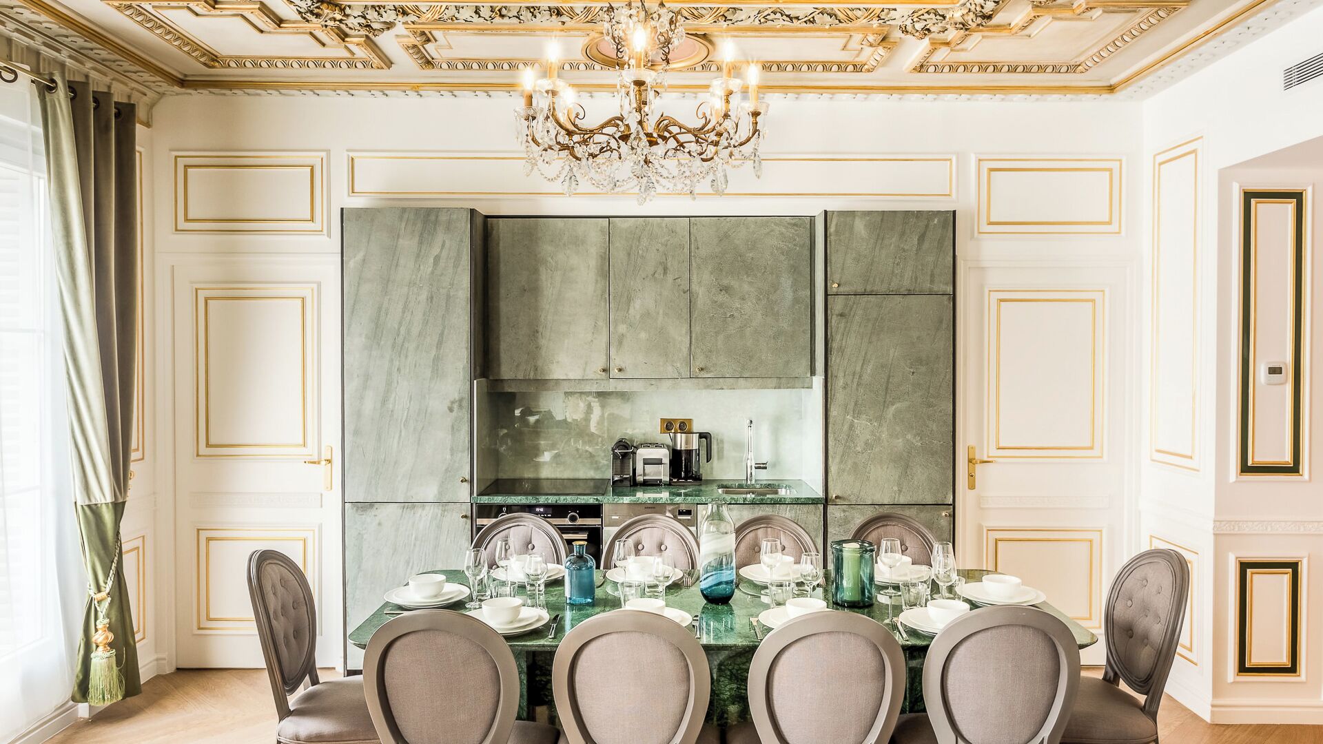 Une salle à manger avec une table en marbre dressée pour six personnes, un lustre et un coin cuisine avec un revêtement mural vert.