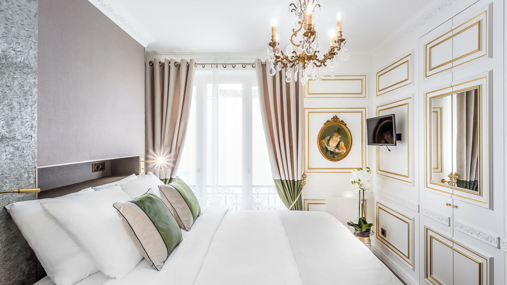 A hotel room with a large bed, a chandelier, a television, and a mirror.