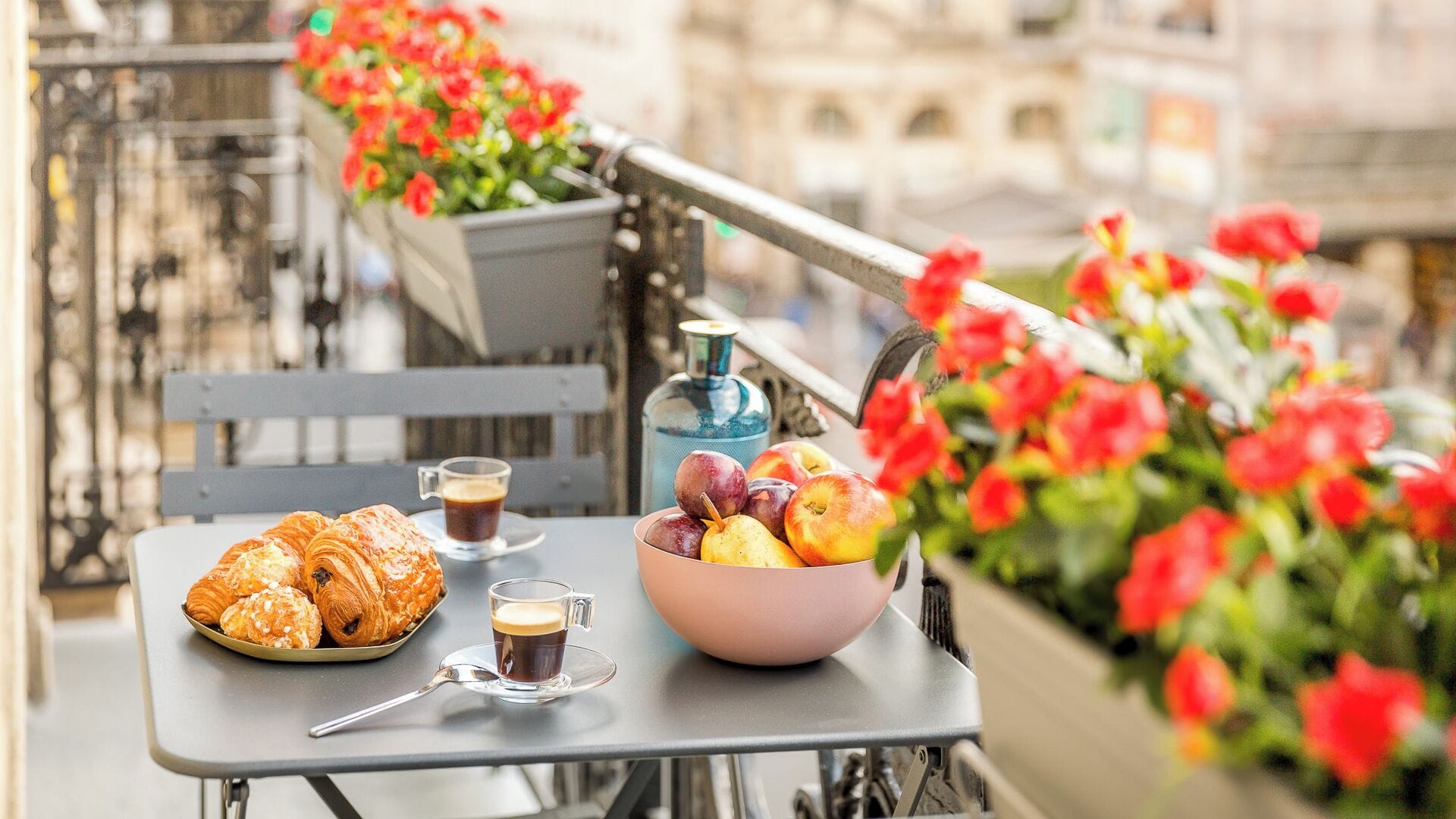 A table with croissants, a coffee cup, and a bowl of fruit on a balcony with red flowers.