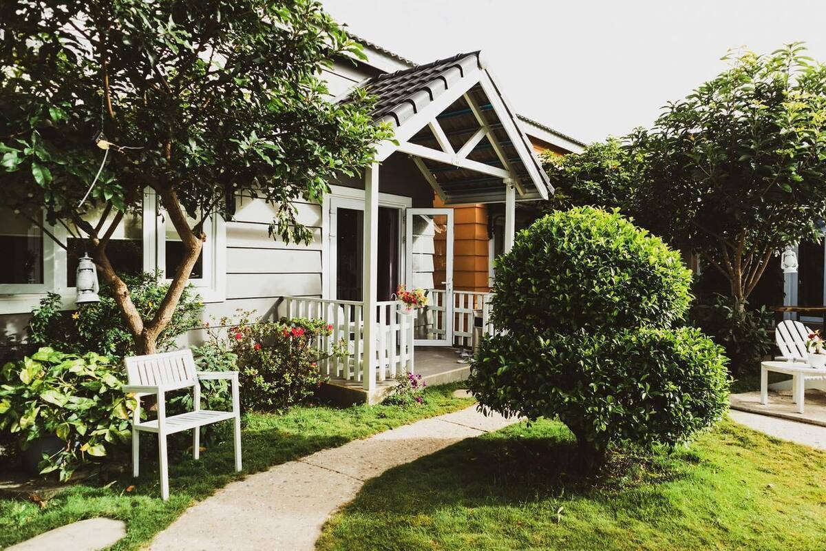 Una casa con porche, un camino y un pequeño jardín con un banco y un árbol.