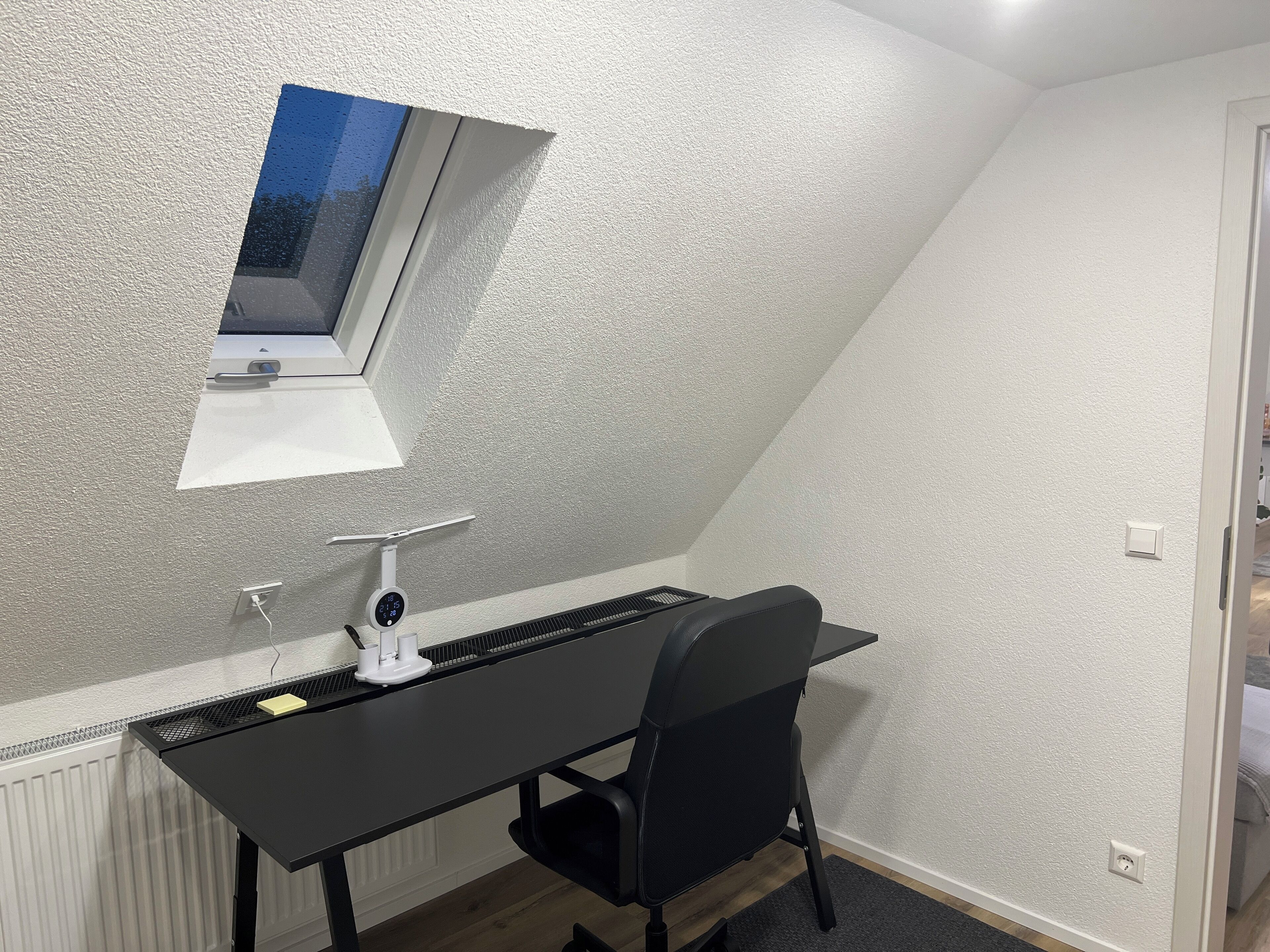 Una stanza piccola e ben illuminata, con soffitto spiovente, una scrivania con sedia, una finestra e un termostato.