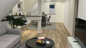 A modern living room with a grey sofa, a round coffee table with a book and a remote control, a dining area with a table and chairs, and a staircase leading to an upper level. - Stylish Apartment for Families, Technicians, and Business Travelers in Horb (Horb am Neckar)