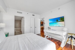 Habitación de hotel con cama, escritorio, silla, televisión y un cuadro de una playa.