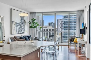 A modern living room with a large window, a sofa, a dining area, and a view of high-rise buildings.