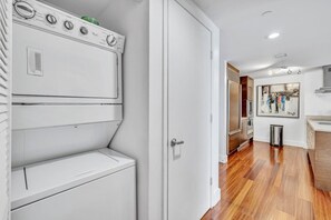 Un lavadero con lavadora y secadora blancas, una puerta que da a un pasillo y una zona de cocina con suelo de madera y un refrigerador de acero inoxidable.