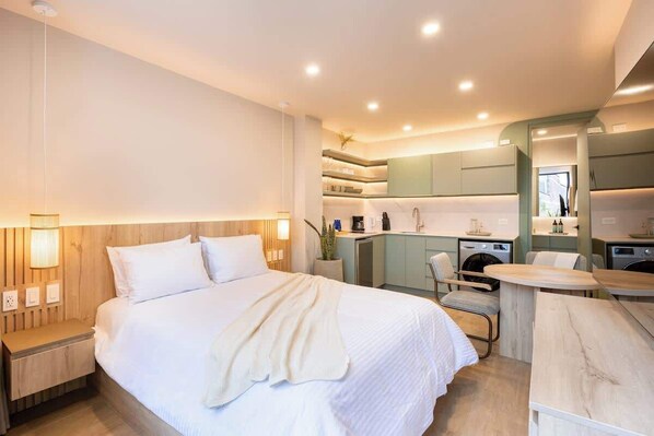 Una habitación de hotel moderna con una cama grande, mesitas de noche, un escritorio pequeño, una silla, una lavadora y una kitchenette.
