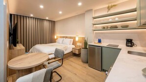Una habitación de hotel moderna con cama, mesita de noche, silla, una pequeña mesa redonda y una zona de kitchenette con un lavabo y armarios.