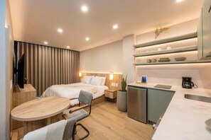 Une chambre d’hôtel moderne comprenant un lit, une table de chevet, une chaise, une petite table ronde et un coin cuisine équipé d’un évier et de placards.