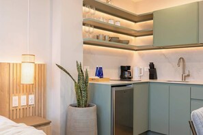 Une cuisine compacte équipée d’un lave-vaisselle intégré, d’une machine à café et d’un évier. On y trouve également une plante en pot et une étagère avec de la verrerie et des assiettes.