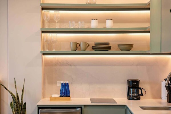 Une cuisine moderne équipée d’un plan de travail, d’une machine à café et d’étagères où sont rangés de la verrerie et de la vaisselle.
