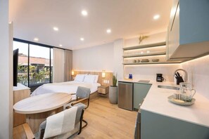 Ein modernes Hotelzimmer mit Bett, Schreibtisch, einer kleinen Küchenzeile und einem großen Fenster mit Blick ins Freie.