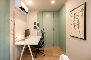 Un petit bureau avec un bureau blanc, une chaise de bureau noire et un écran d’ordinateur. Un tableau abstrait encadré est accroché au mur, et un appareil de climatisation est fixé au mur.