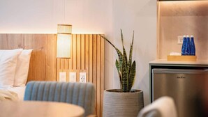Una habitación de hotel moderna con cabecera de madera, lámpara de noche, planta en maceta, minibar y cama con ropa de cama blanca.