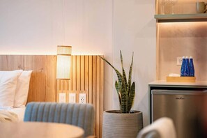Una habitación de hotel moderna con cabecera de madera, lámpara de noche, planta en maceta, minibar y cama con ropa de cama blanca.