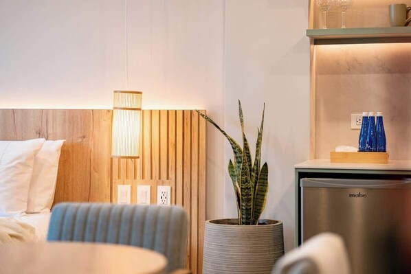 Ein modernes Hotelzimmer mit einem Holz-Kopfteil, einer Nachttischlampe, einem Topf mit Pflanze, einer Minibar und einem Bett mit weißen Bettwäsche.