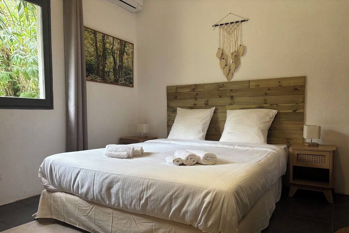 A neatly made bed with white linens and rolled towels, a wooden headboard, a wall-mounted macrame piece, and a small bedside table with a lamp.