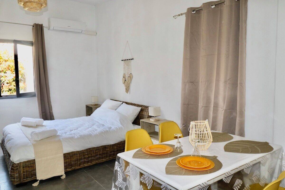 A room with a bed, a dining table with yellow chairs, and a window with curtains.