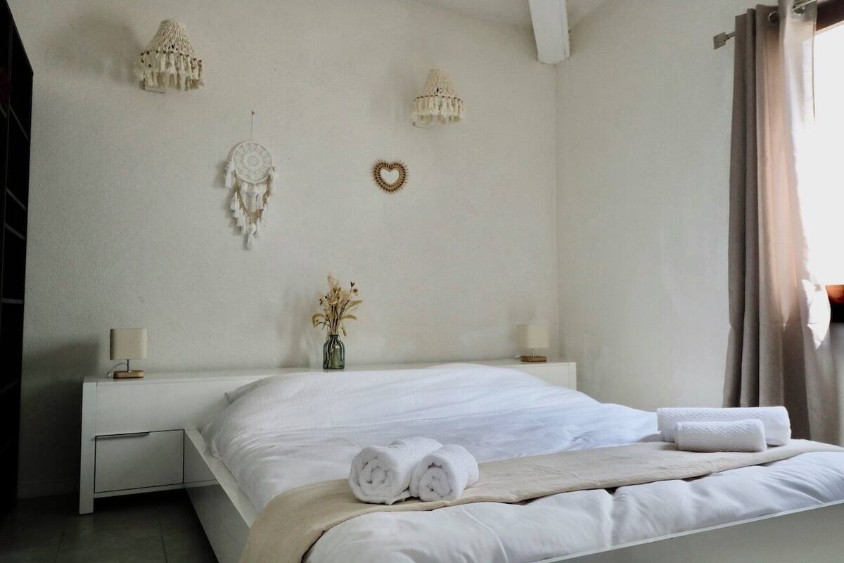 A neatly made bed with rolled towels, a bedside table with a lamp, a dreamcatcher, and a heart-shaped decoration on a white wall.