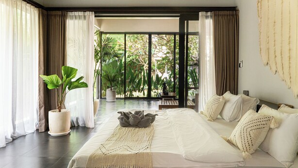 A spacious bedroom with a large bed, a potted plant, and a view of greenery through the window.