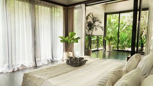 A large bed with a white bedspread and pillows, a potted plant, and a view of lush greenery through large windows.