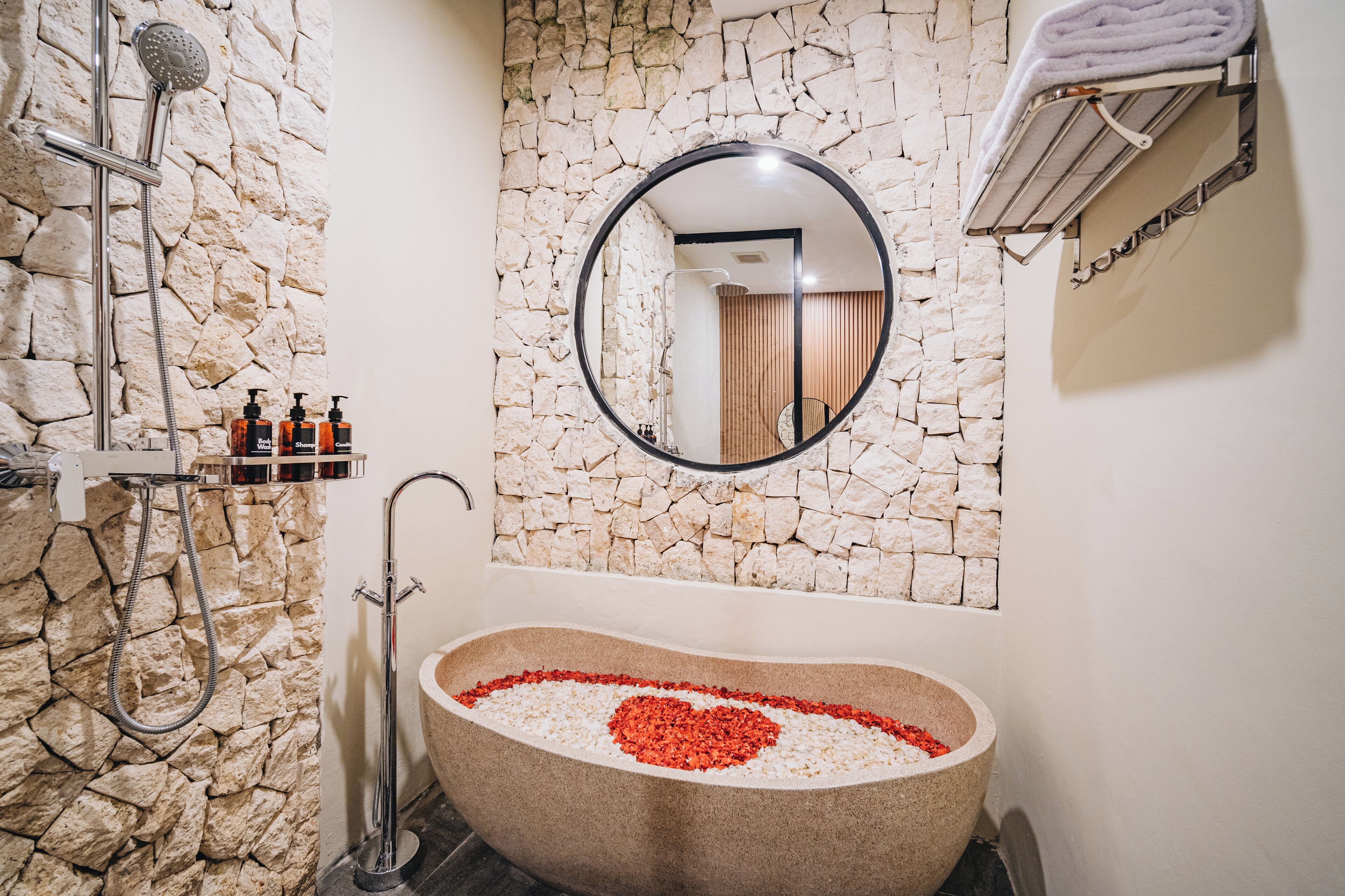 A bathroom with a stone wall, a freestanding bathtub with a heart-shaped arrangement of red and white pebbles, a round mirror, and a towel rack.
