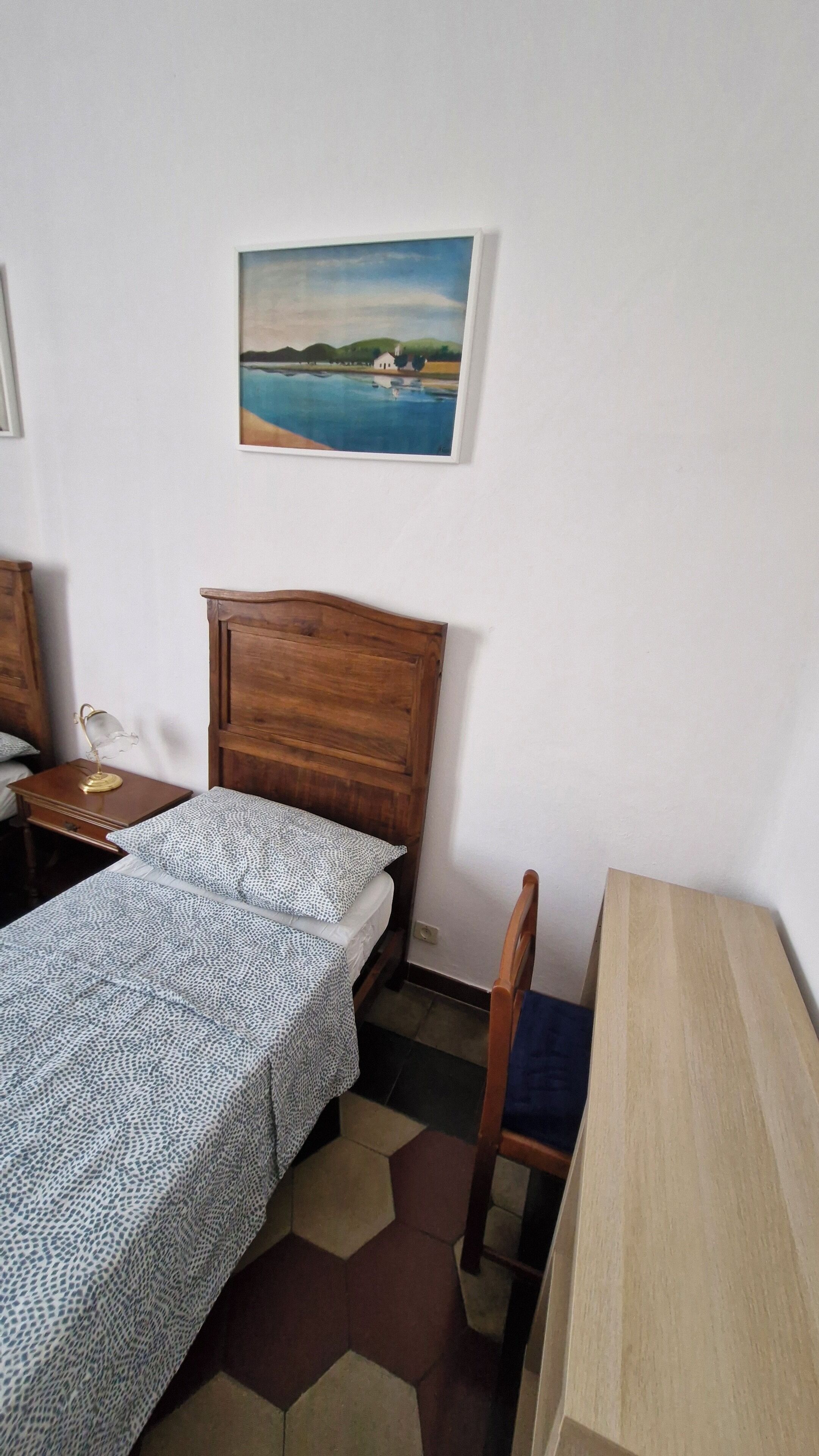 Quarto com cama de madeira, uma mesinha de cabeceira com abajur, uma cadeira e uma escrivaninha de madeira.