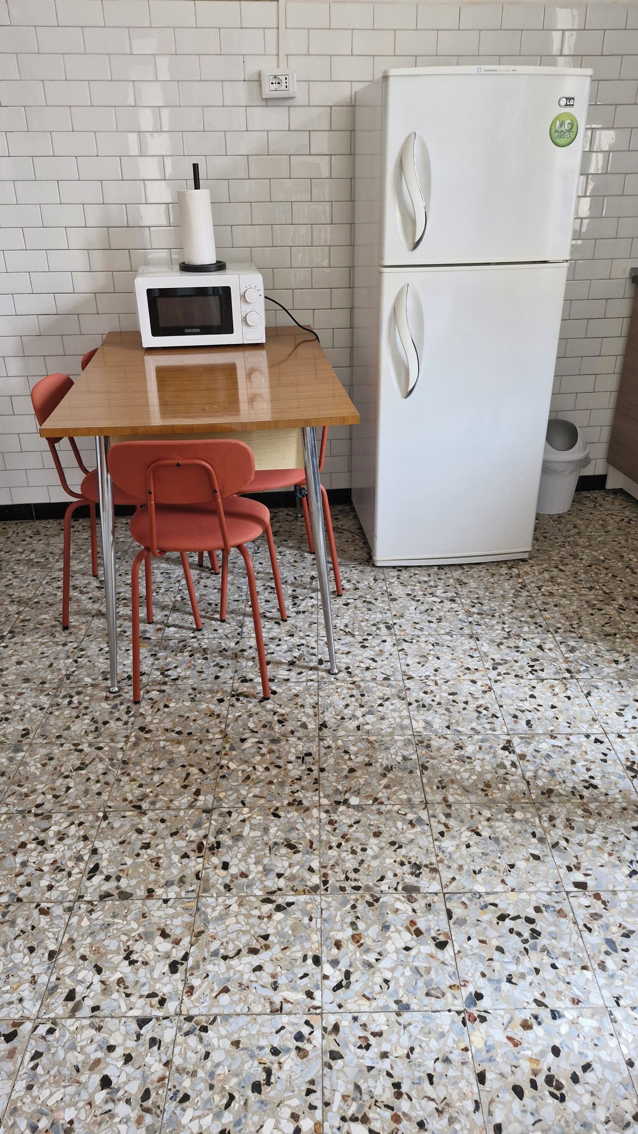 Eine Küche mit einem weißen Kühlschrank, einer Mikrowelle und einem Tisch mit roten Stühlen. Der Boden weist ein gemustertes Fliesenmuster auf.