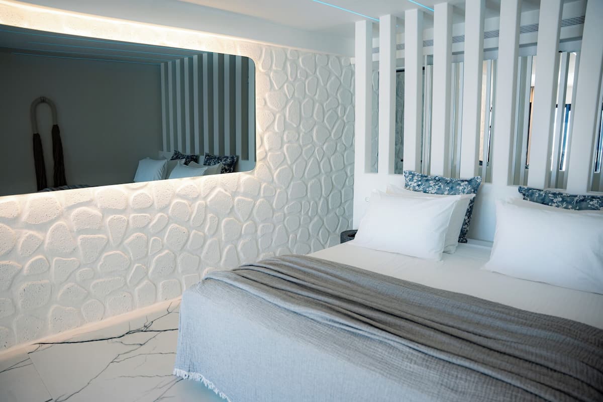 Un dormitorio moderno con una pared con textura de piedra, una cama grande con una manta gris y un espejo que refleja la habitación.