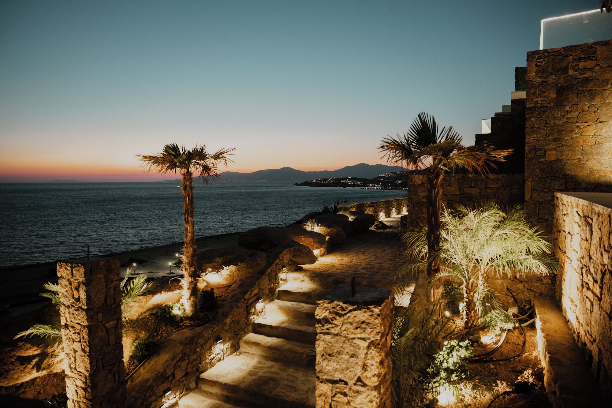A coastal pathway with stone steps and palm trees, leading to a beach with a sunset view.