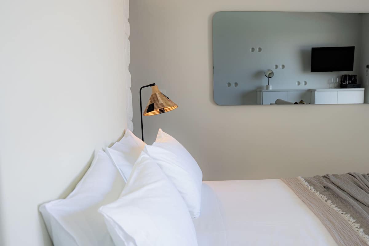 Una cama bien tendida con sábanas blancas, una lámpara con pantalla tejida, un espejo grande que refleja un televisor y un mueble blanco con cafetera.