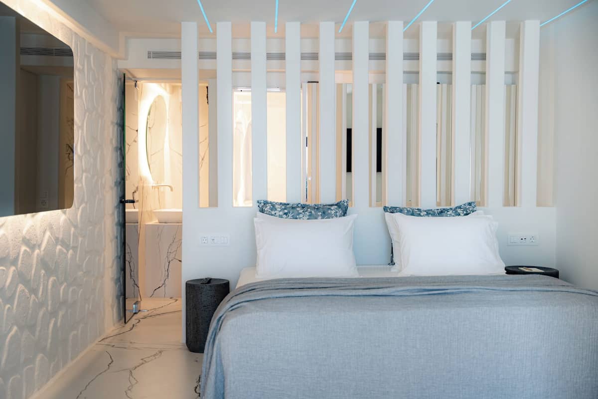 Un dormitorio moderno con cama, mesita de noche, televisor empotrado en la pared y ventana con persianas verticales.