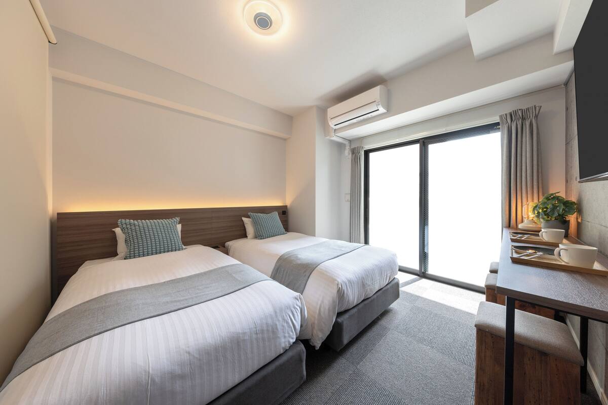 Quarto de hotel com duas camas, uma escrivaninha com cadeira e uma janela grande com cortinas.