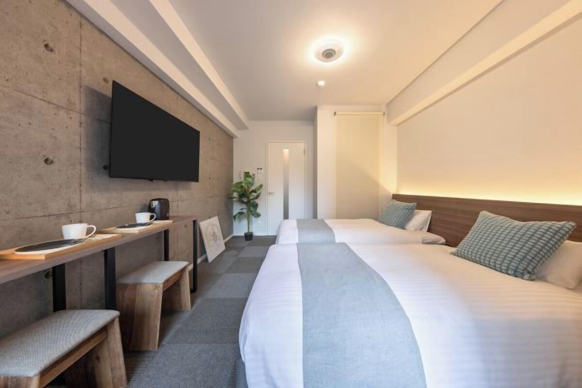 A hotel room with a bed, a desk with a chair, a flat-screen TV mounted on the wall, and a potted plant.