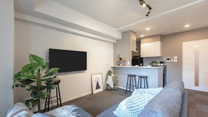 A modern living room with a grey sofa, a flat-screen TV mounted on the wall, a small kitchen area with bar stools, and a large potted plant.