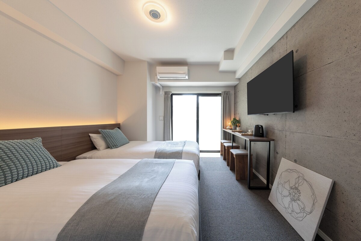 A modern hotel room with two beds, a flat-screen TV mounted on the wall, and a large window providing a view of the outdoors.