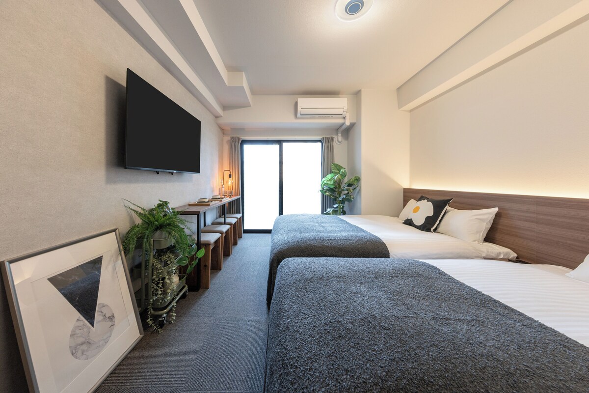 A hotel room with two beds, a TV mounted on the wall, a desk with a lamp, and a large window.