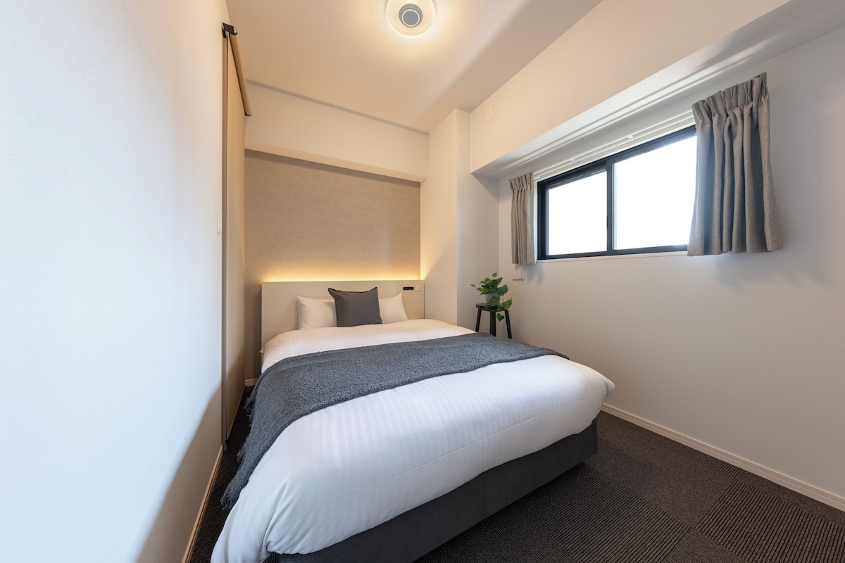 A neatly arranged hotel room with a large bed, a window with curtains, a small table with a plant, and a wall-mounted light fixture.