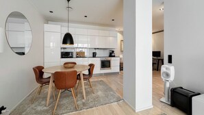 A modern kitchen with white cabinets, a dining area with a round table and chairs, and a visible TV in the adjacent room.