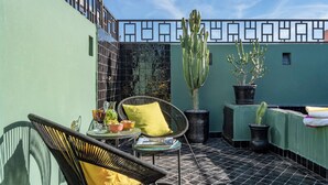 Una terrazza panoramica con parete verde, pavimento piastrellato, sedie in vimini con cuscini gialli, un tavolo con bevande e piante in vaso.