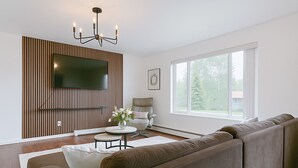Ein modernes Wohnzimmer mit einem Ecksofa, einem Flachbildfernseher an einer Wand aus Holzpaneelen, einem runden Couchtisch mit Blumen in einer Vase und einem großen Fenster mit Blick auf Bäume.